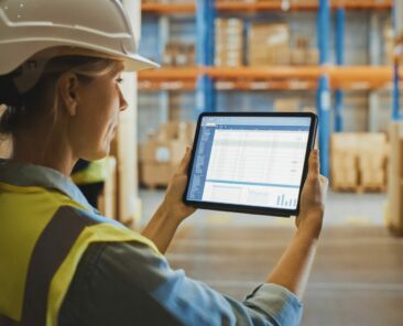 Professional Female Worker Wearing Hard Hat Uses Digital Tablet Computer with Screen Showing Inventory Checking Software in the Retail Warehouse full of Shelves with Goods. Over the Shoulder View