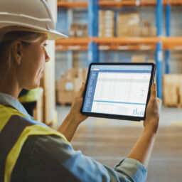 Professional Female Worker Wearing Hard Hat Uses Digital Tablet Computer with Screen Showing Inventory Checking Software in the Retail Warehouse full of Shelves with Goods. Over the Shoulder View